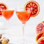 Blood Orange whiskey sour with a blood orange slice as garnish. Another drink is in the background and there are blood oranges and ginger on the countertop.