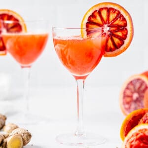 Blood Orange whiskey sour with a blood orange slice as garnish. Another drink is in the background and there are blood oranges and ginger on the countertop.