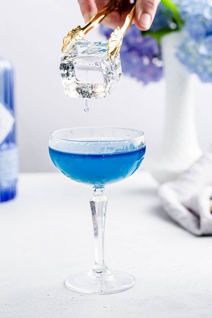 holding dripping clear ice over blue cocktail in a coupe glass