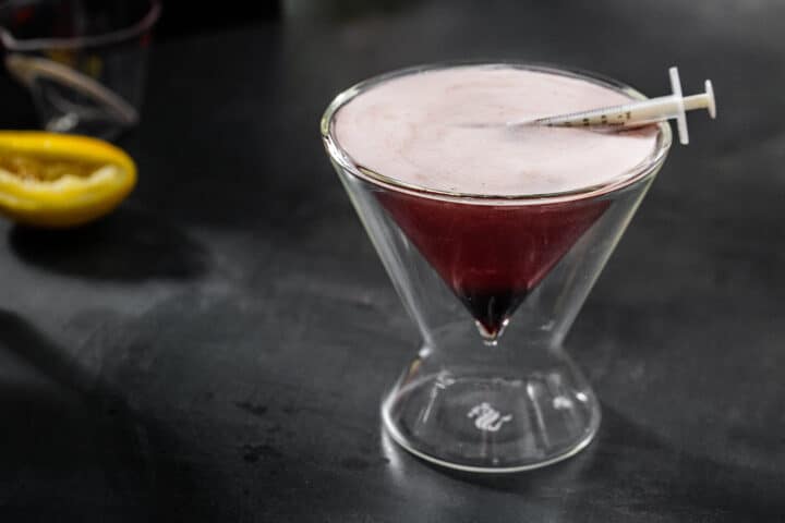 Horizontal image of bloody shiraz sour Halloween gin cocktail, with medicine dropper garnish and a squeezed lemon in the background.