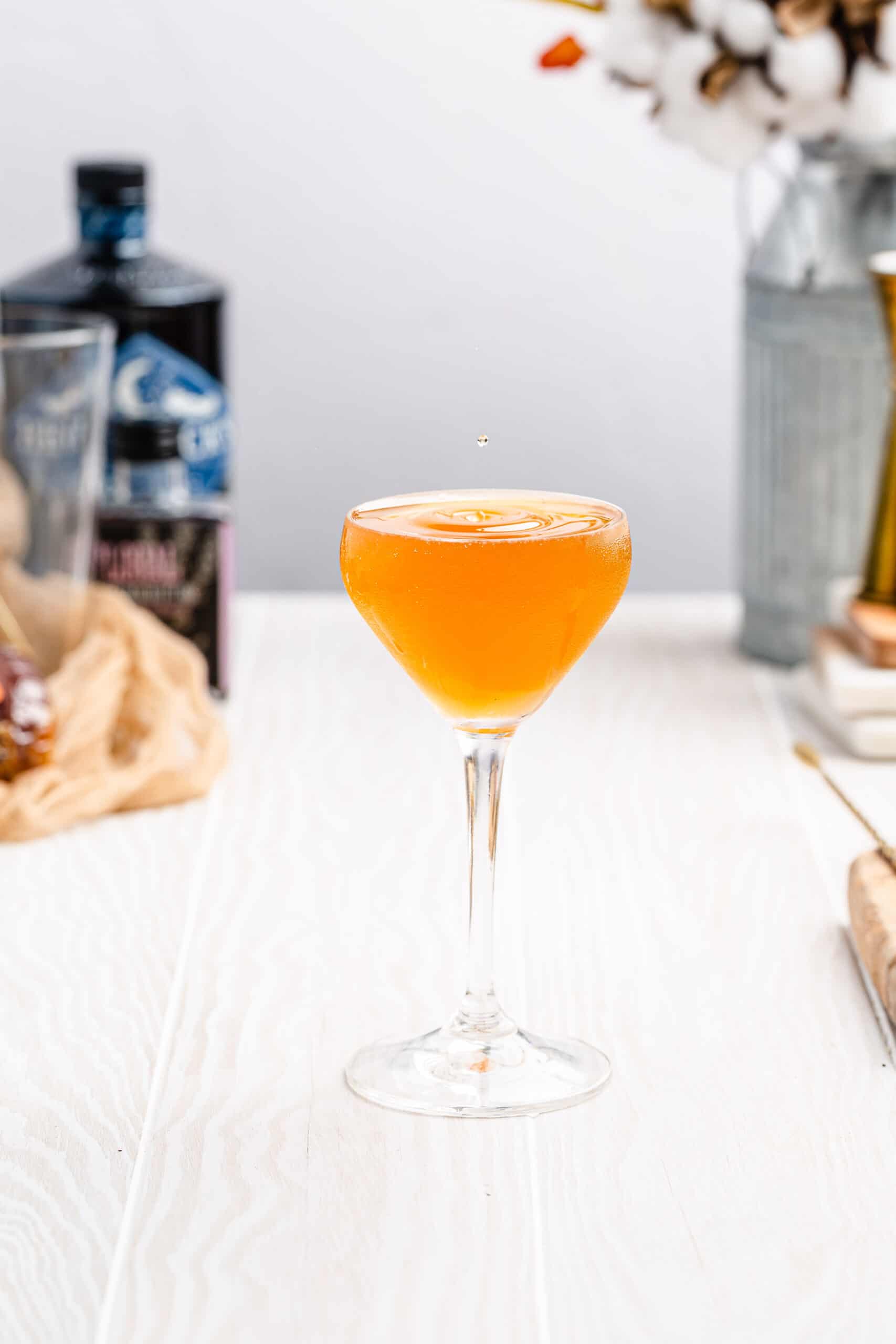 vertical image of bees knees cocktail in a stemmed cocktail glass with ingredients in the background