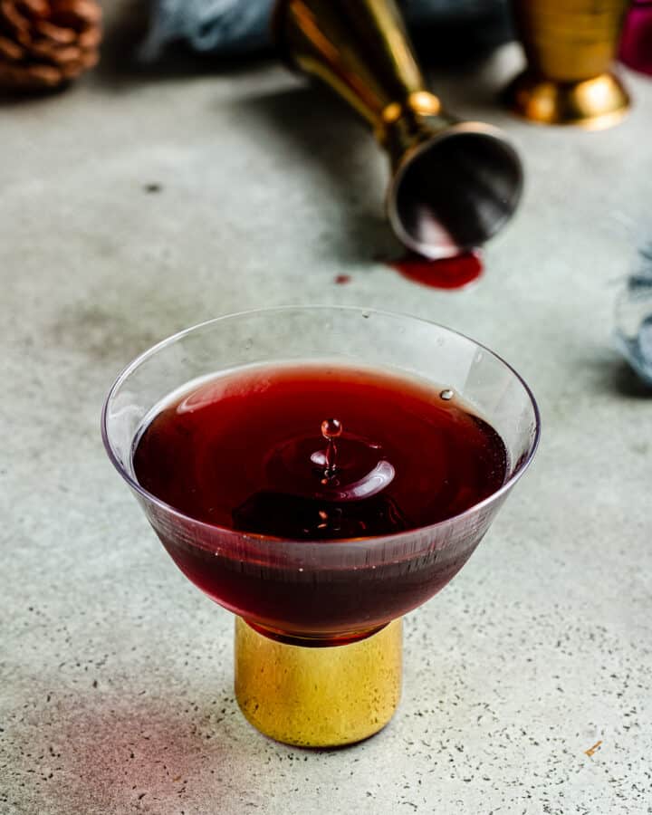 dark red cocktail in glass with gold stem and jigger in the background