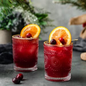 two cranberry and orange juice mocktails surrounded by cranberries and evergreens