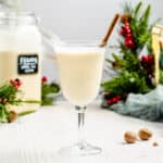 side view of glass goblet filled with eggnog and a cinnamon stick with christmas decor in the background