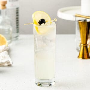 Side view of a classic Tom Collins in a skinny glass with a gold jigger in the background