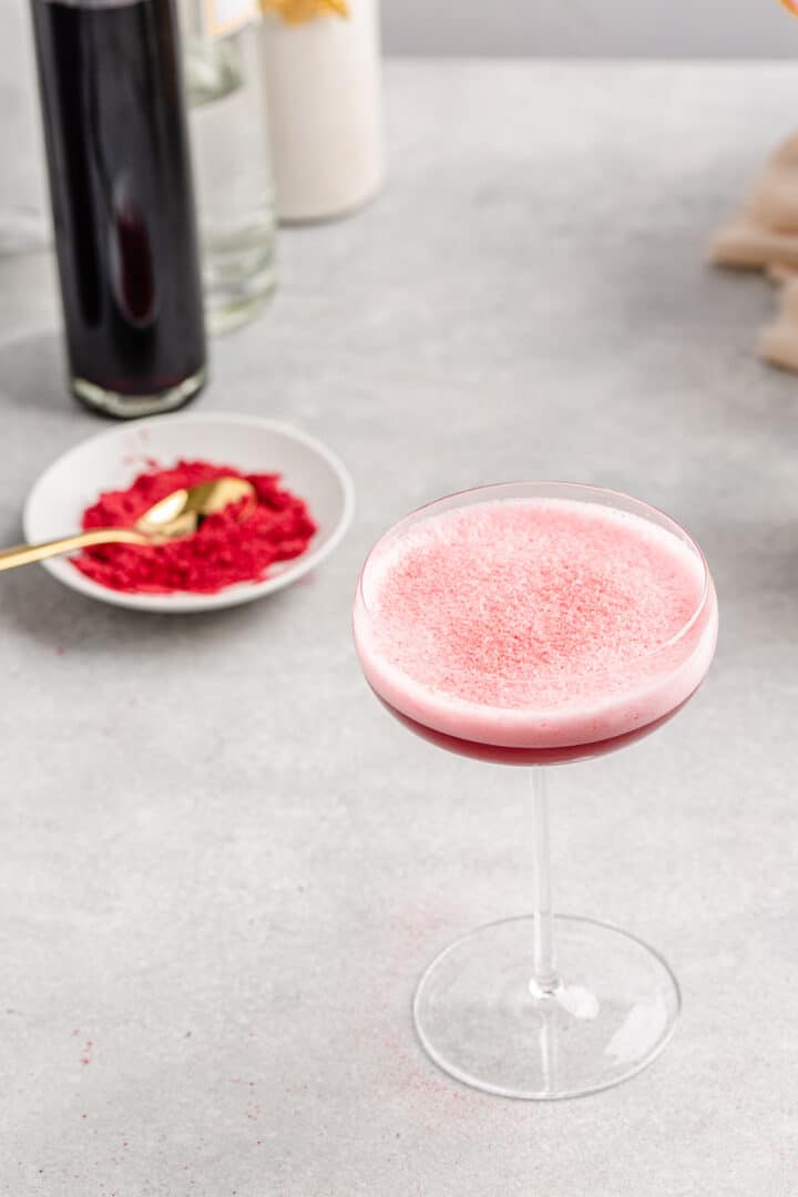 Top view of pink foamy cocktail with red powder in a bowl and bottles in the background.