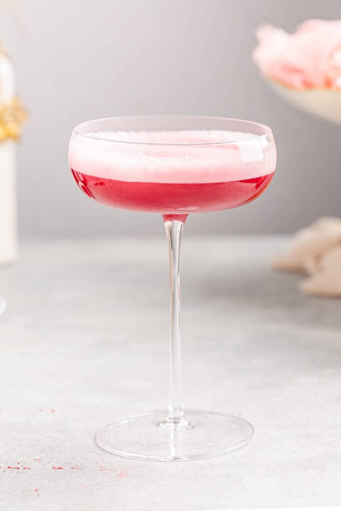 Side view of a hibiscus vodka cocktail. The drink is bright pink on the bottom with light pink foam on top. a dish with pink roses is in the background giving it a romantic feel.