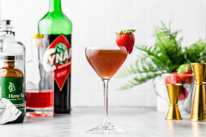 Side view of a cocktail with a strawberry garnish and ingredients and jiggers in the background.