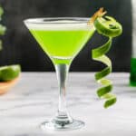 Side view of a Martini glass with a bright green Midori Sour in it and a long curly strip of lime zest clipped to the glass.