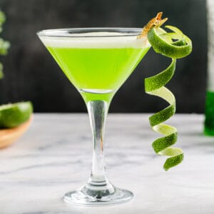 Side view of a Martini glass with a bright green Midori Sour in it and a long curly strip of lime zest clipped to the glass.