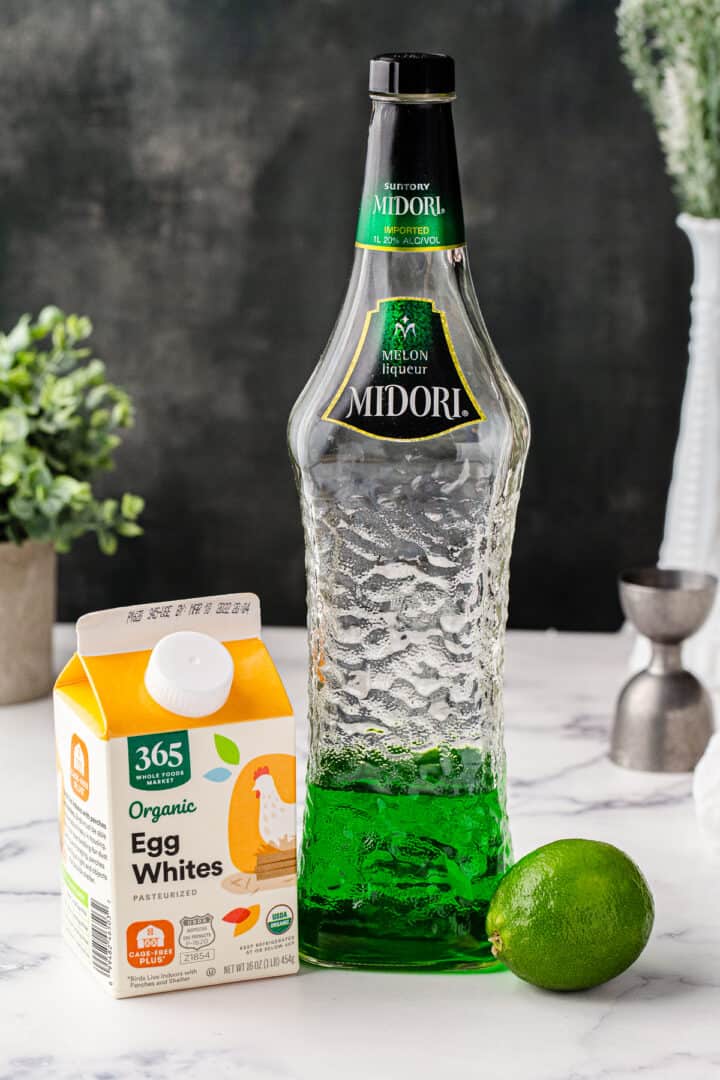 A bottle of Midori next to egg whites and a lime on a marble countertop.
