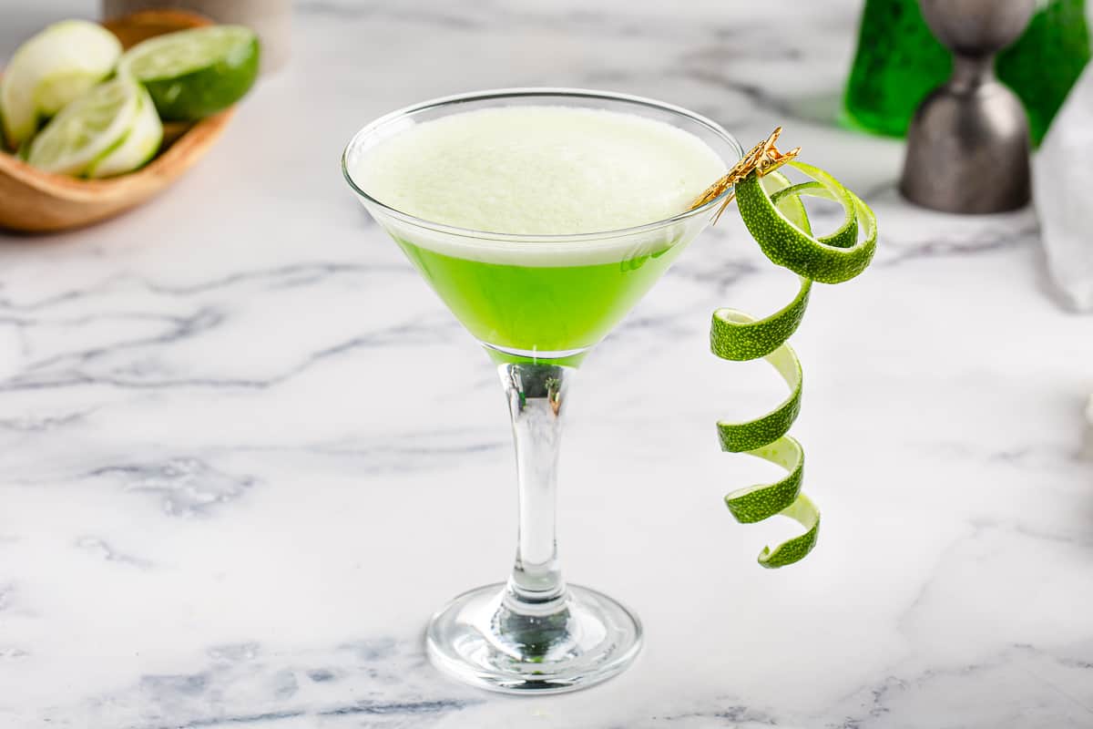 Midori Sour with a curly lime zest garnish on a marble countertop.
