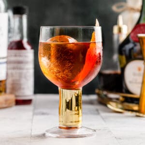 Side view of the Vieux Carre cocktail with an orange peel garnish and bottles in the background.
