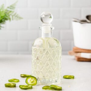 Side view of a glass bottle with jalapeno syrup in it and jalapeno slices around it and cooking equipment in the background.