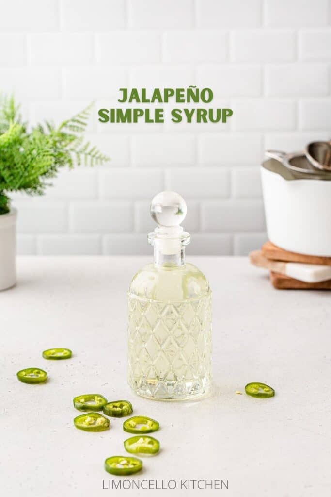 Side view of a glass bottle with jalapeno syrup in it and jalapeno slices around it and a white pot for cooking in the background.