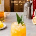 Cocktail in a lowball glass with pineapple frond garnish, and bar tools and ingredients in the background, with the text Pineapple Whiskey Smash cocktail above it.