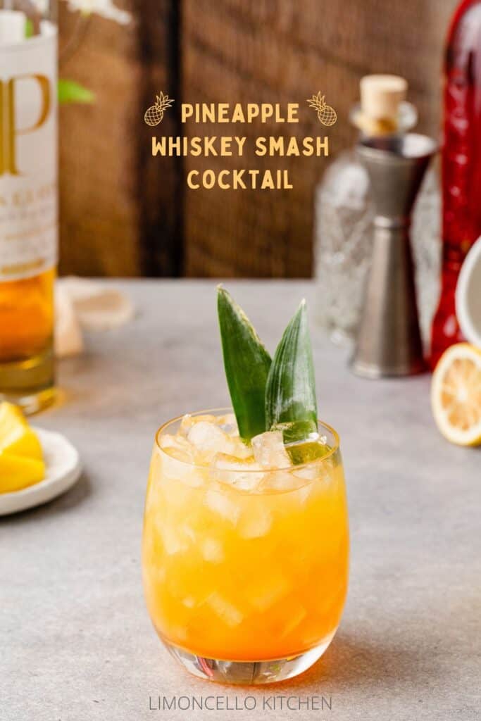 Cocktail in a lowball glass with pineapple frond garnish, and bar tools and ingredients in the background, with the text Pineapple Whiskey Smash cocktail above it.