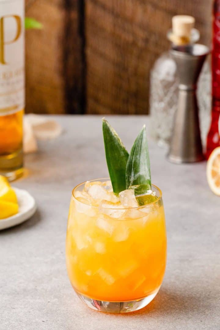 Pineapple Whiskey Smash cocktail in a lowball glass with nugget ice and a pineapple frond as a garnish. Ingredients and bar tools are in the background.