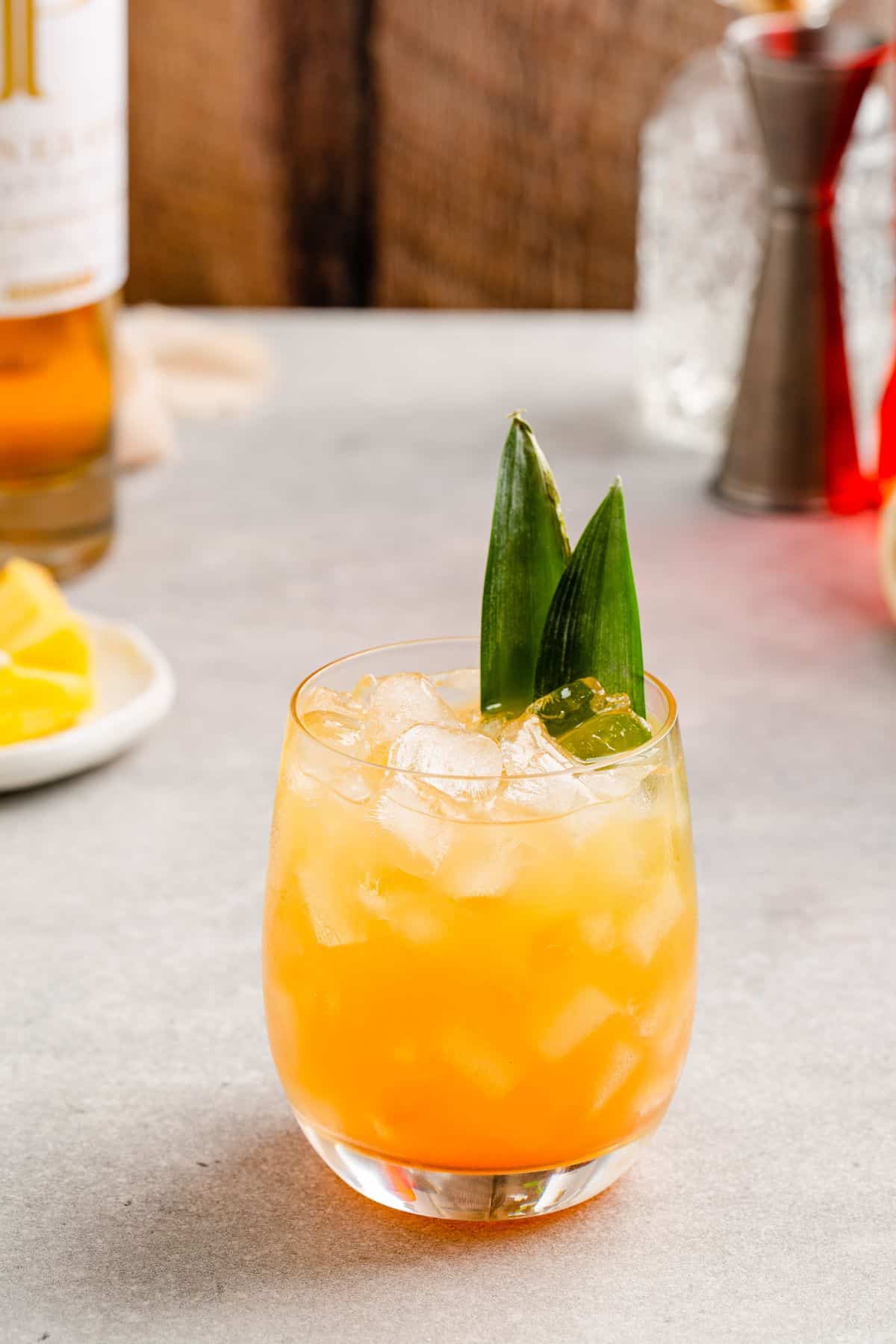 Pineapple Whiskey Smash cocktail in a lowball glass with nugget ice and a pineapple frond as a garnish. Ingredients and bar tools are in the background.