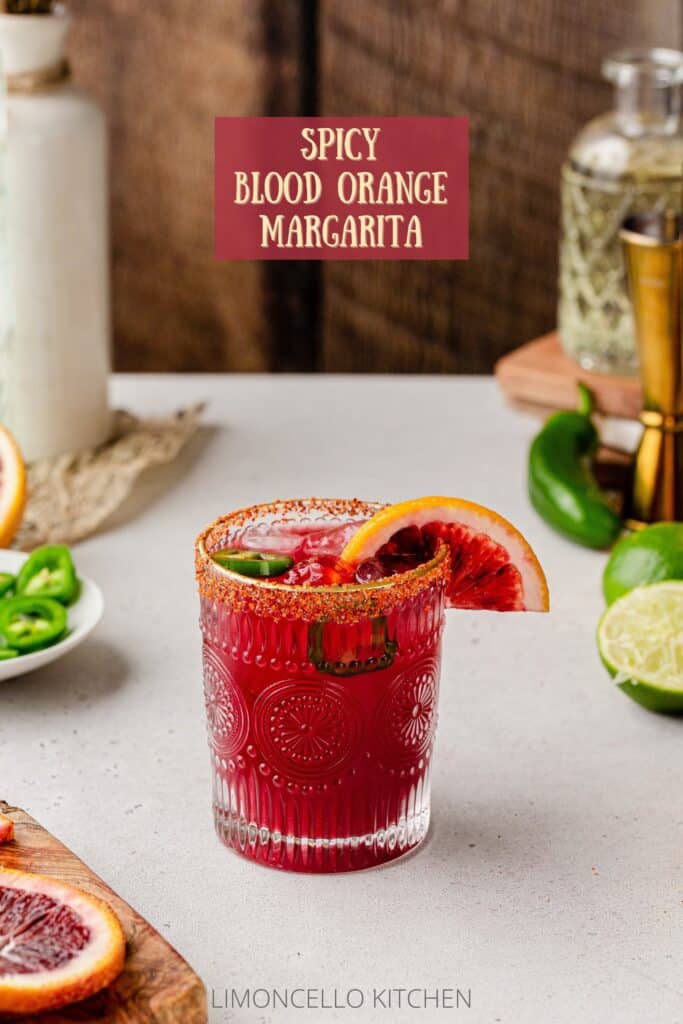 Spicy Blood Orange Margarita with a jalapeño and blood orange garnish on a countertop with fresh ingredients and bar tools in the background.