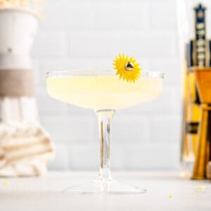 Side view of the Sunflower cocktail in a coupe glass with ingredients and tools in the background.