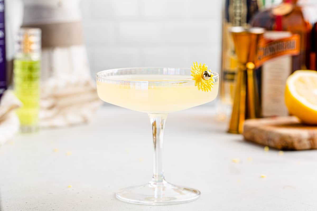 Side view of a yellow colored Sunflower cocktail in a wide coupe glass with cocktail tools and ingredients in the background.