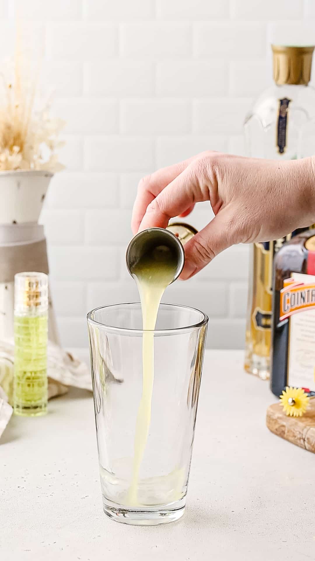 Hand pouring lemon juice from a jigger into a glass cocktail shaker.