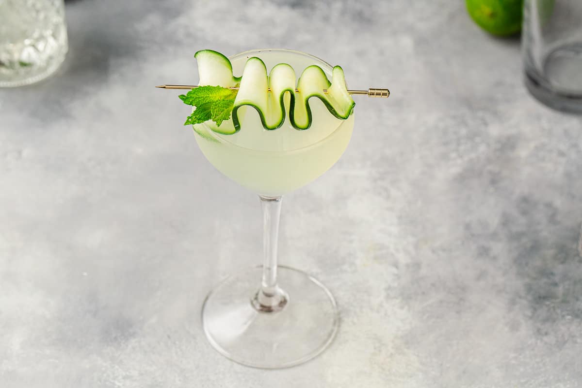 Close up view of the Eastside cocktail on a gray countertop with a zigzag cucumber strip garnish and a sprig of mint.