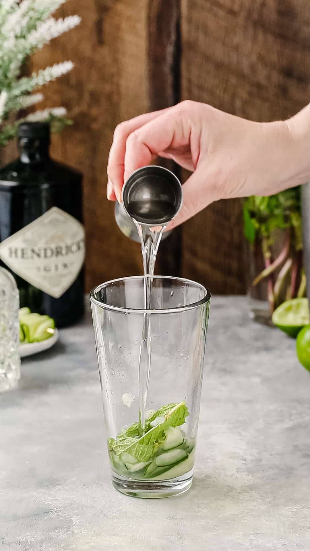 Hand pouring clear liquid from a silver jigger into a cocktail shaker.
