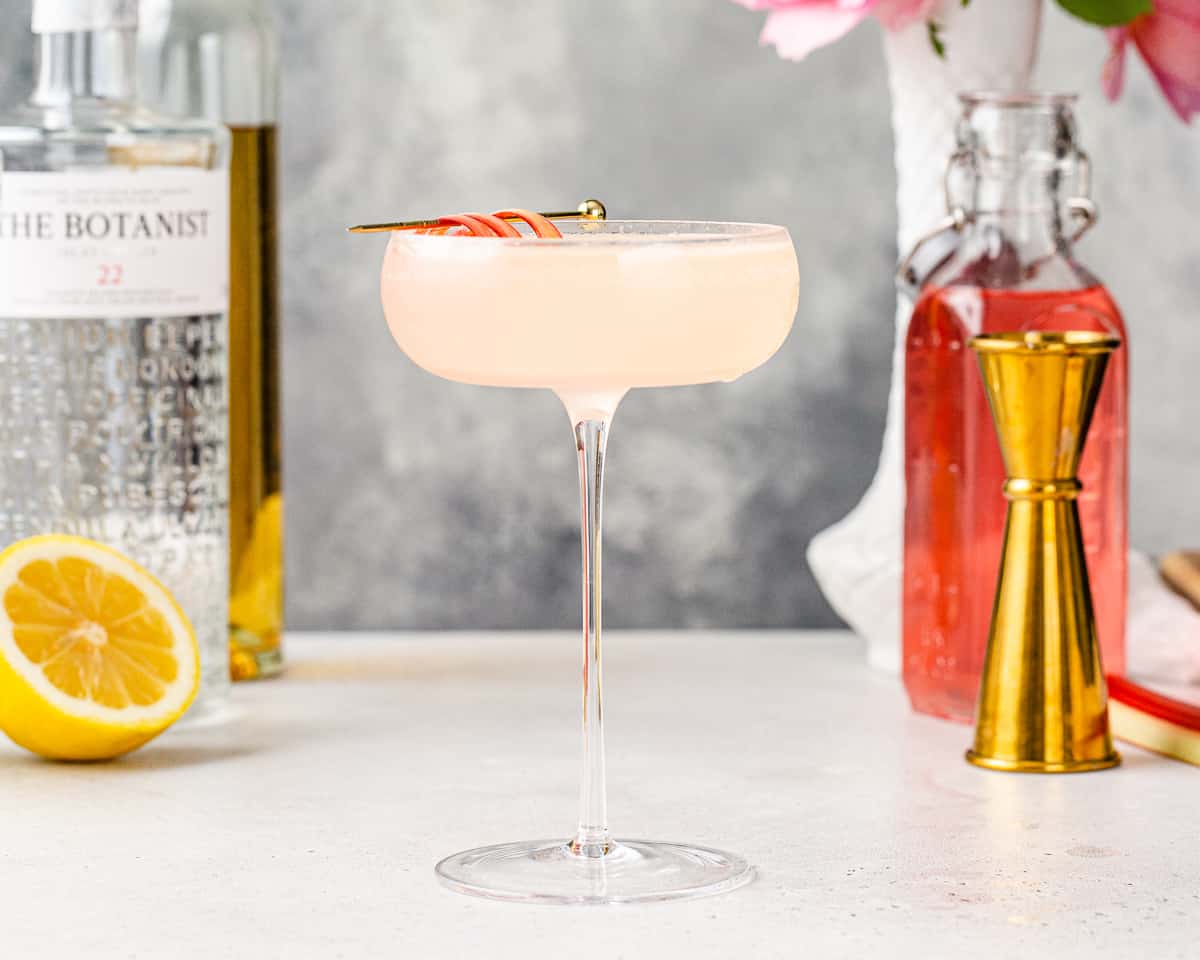 Side view of Rhubarb Gin cocktail in an elegant stemmed glass with bar tools, a ingredients and flowers in the background
