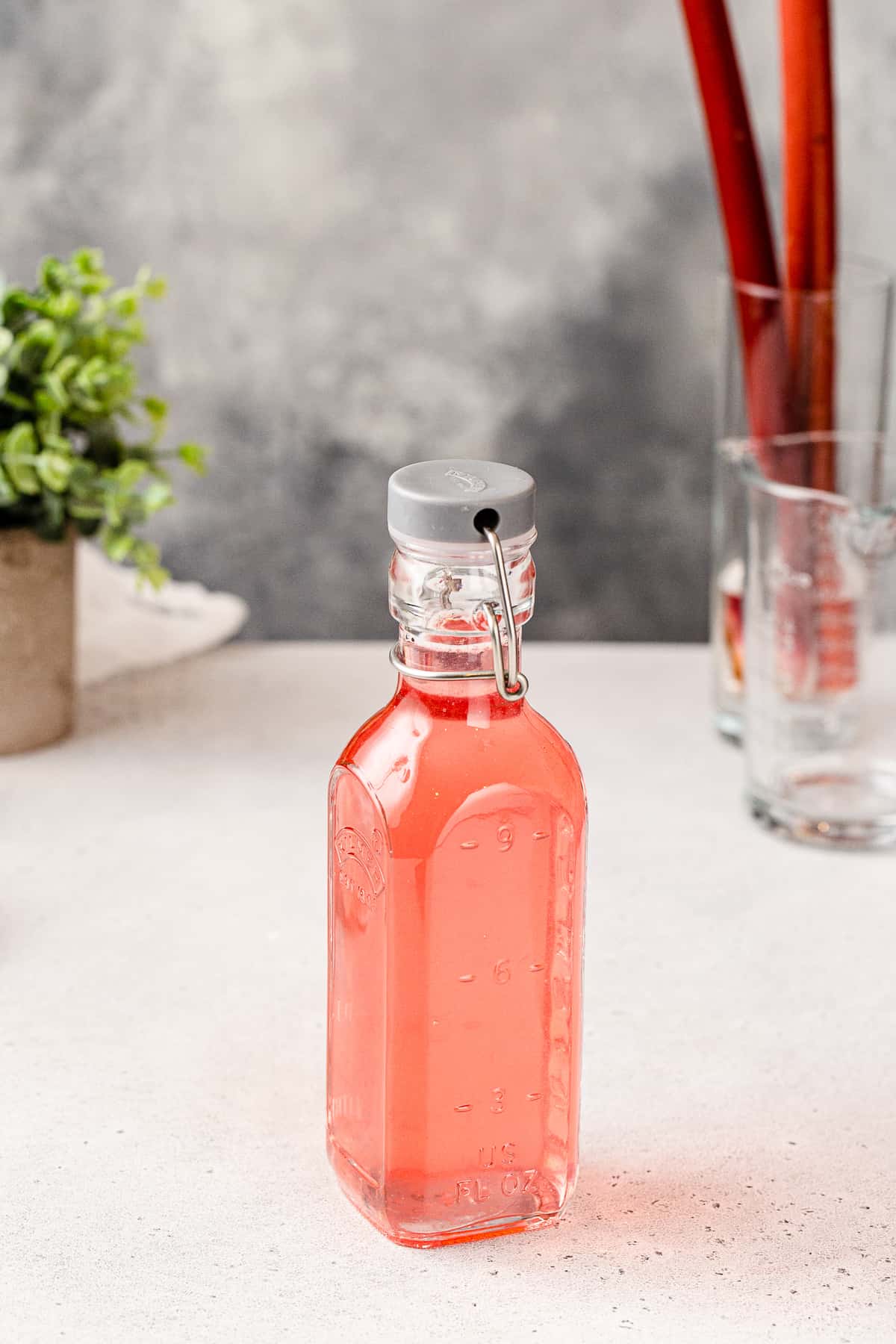 Glass bottle filled with pink rhubarb syrup with greenery and stalks of rhubarb in the background.