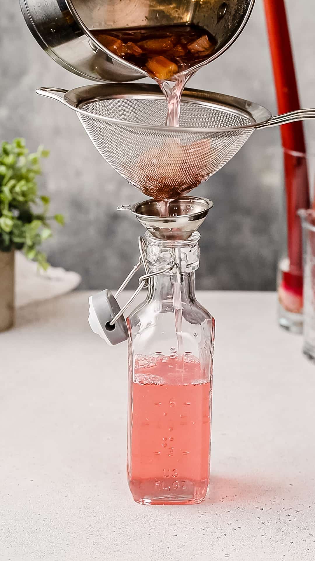 Saucepan with liquid and rhubarb pieces being poured through a strainer and funnel into a glass bottle.