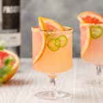 Two Spicy Paloma cocktails on a white wooden countertop with tequila and grapefruit in the background.