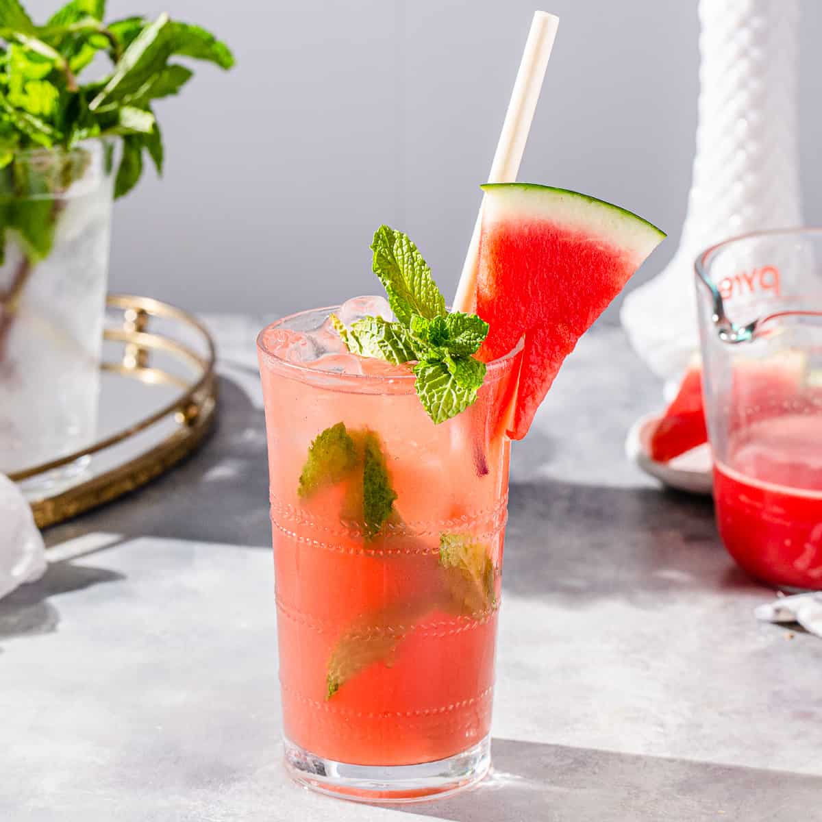 Watermelon mojito cocktail on a countertop with mint and a watermelon slice as a garnish, and more mint and watermelon juice in the background.
