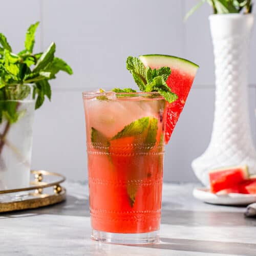 Side view of a watermelon mojito cocktail with mint and a watermelon slice as a garnish, and more mint and watermelon in the background.
