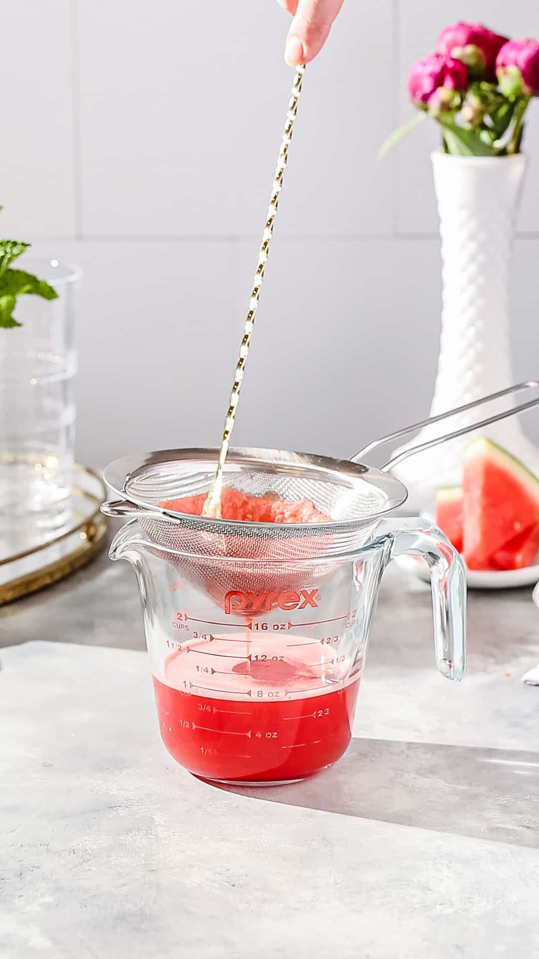 Stirring watermelon puree in a fine mesh strainer using a long bar spoon while the juice drains into a container.
