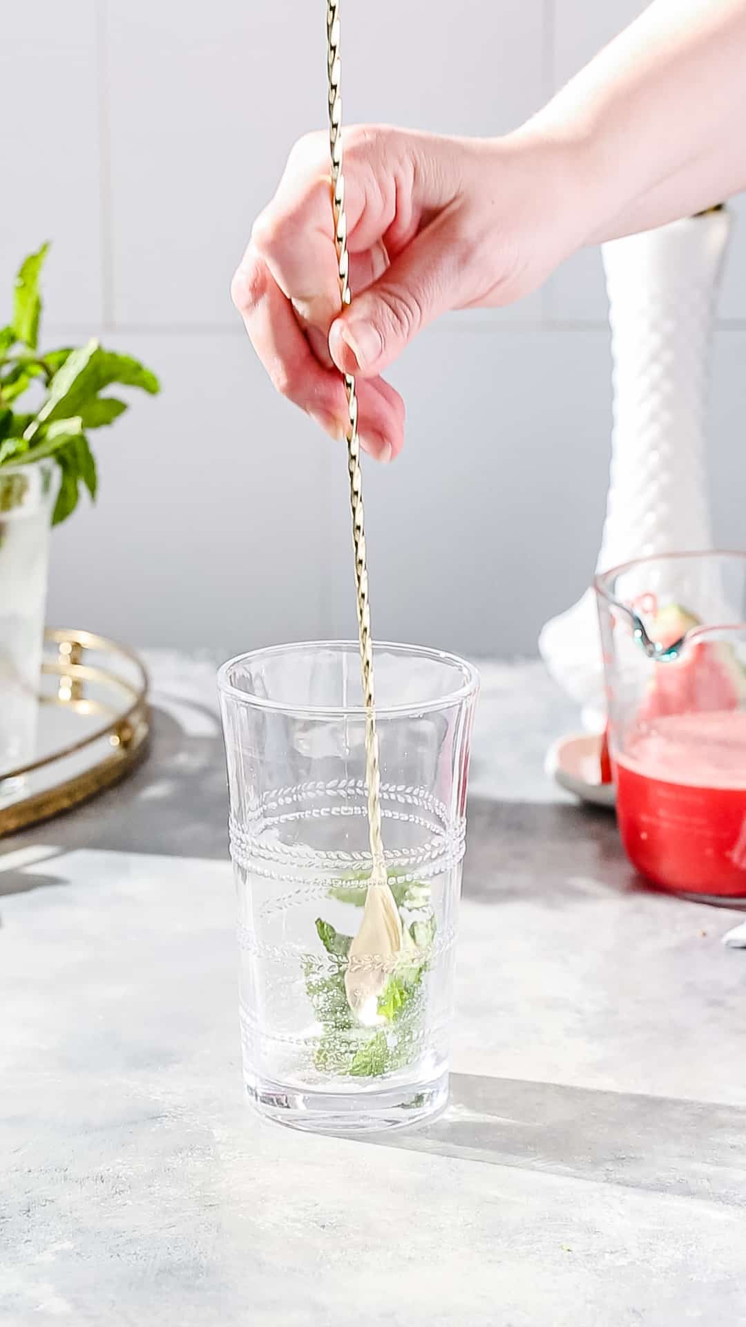 Hand using a long bar spoon to stir mint and sugar together in a cocktail glass.