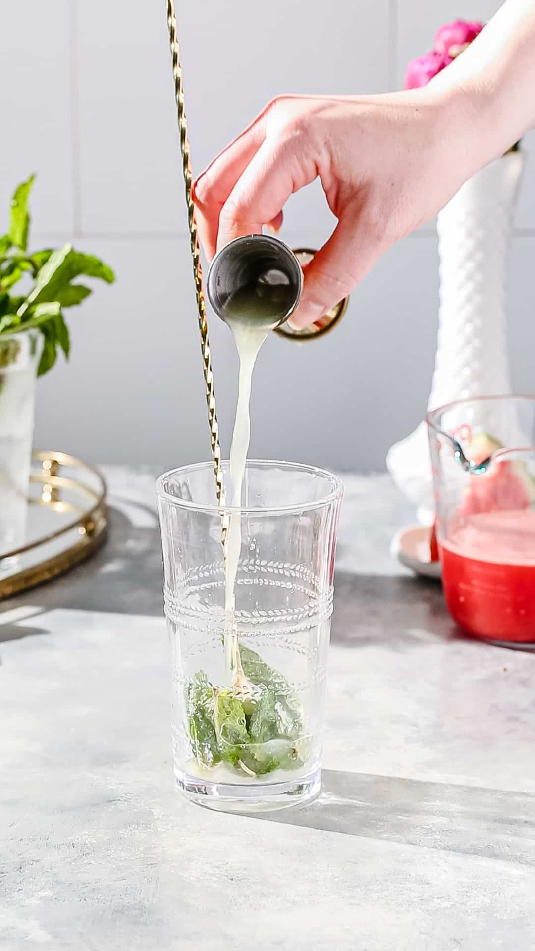 Hand using a jigger to add lime juice to a cocktail glass filled with sugar and mint.