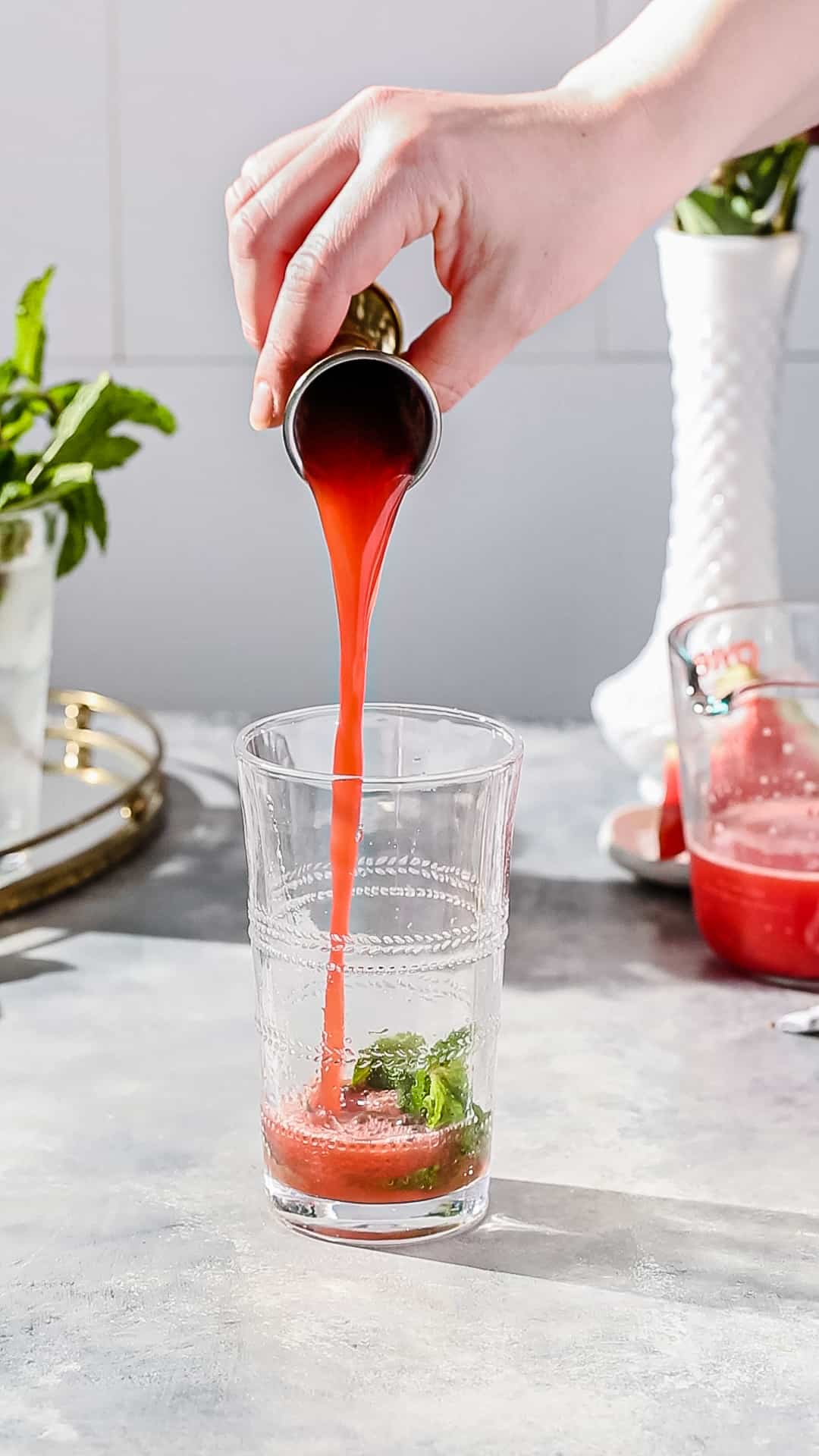Hand using a jigger to add watermelon juice to a cocktail glass filled with mint, sugar and lime juice.