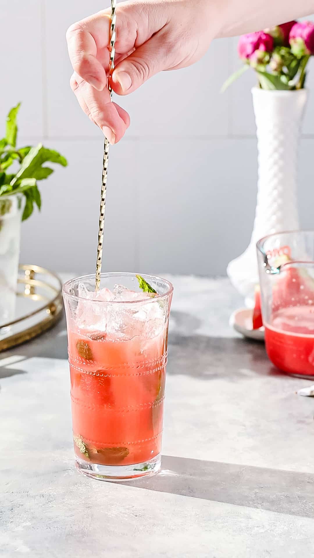 Hand using a long bar spoon to stir a cocktail in a glass.