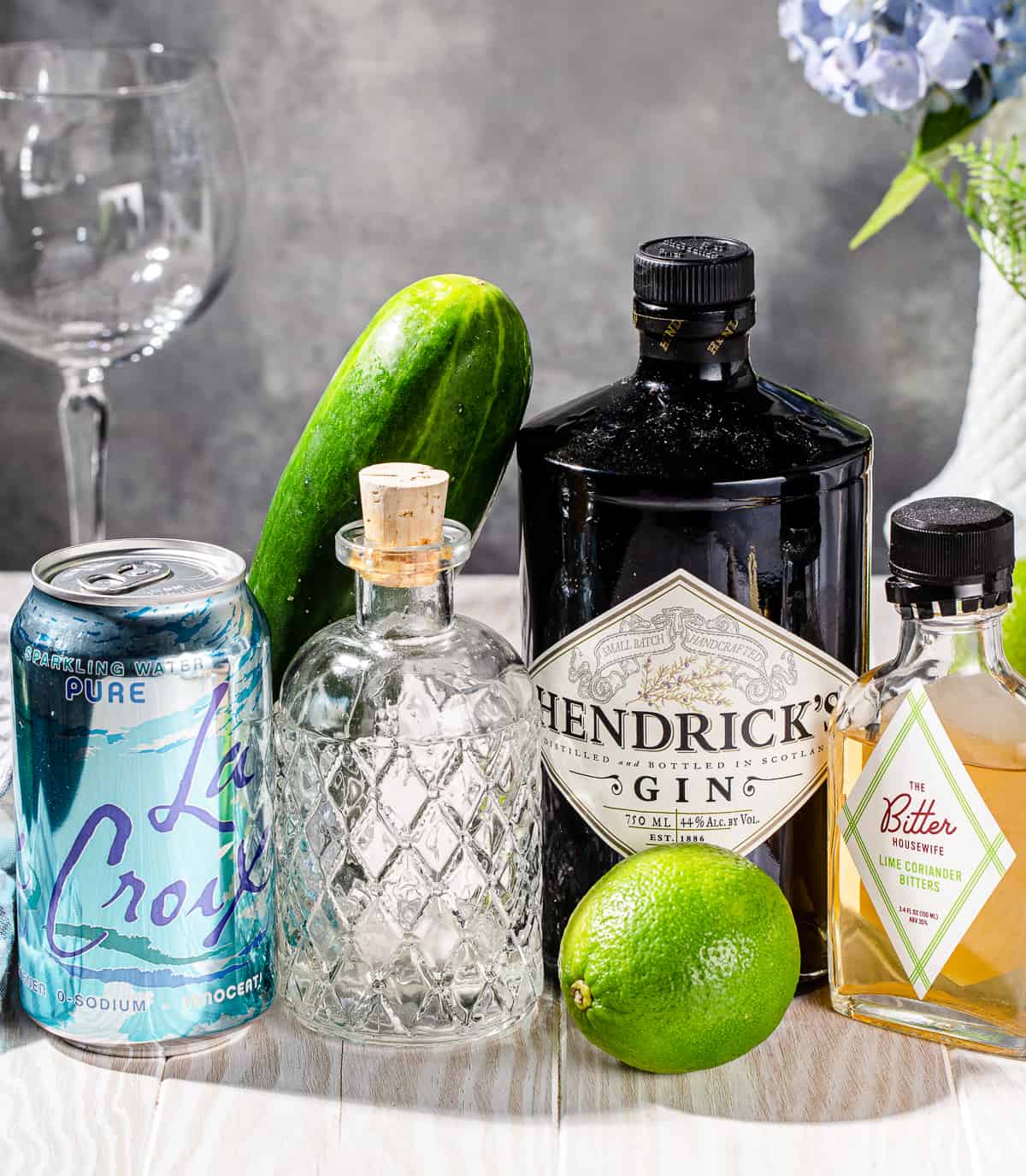 Ingredients to make a Cucumber gin spritzer cocktail set out on a white wood countertop. 