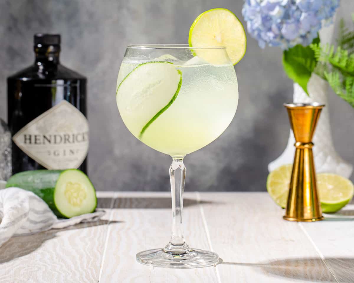 Side view of a Cucumber Gin Spritzer cocktail on a white wood countertop. The drink is in a tall stemmed glass with a round bowl. It is light green and semi transluscent, and the glass has a strip of cucumber inside and a lime wheel on the rim. In the background are bar tools and hydrangea flowers along with ingredients.
