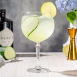 Square photo showing a side view of a Cucumber Gin Spritzer cocktail on a white wood countertop. The drink is in a tall stemmed glass with a round bowl. It is light green and semi transluscent, and the glass has a strip of cucumber inside and a lime wheel on the rim. In the background are bar tools and hydrangea flowers along with ingredients.
