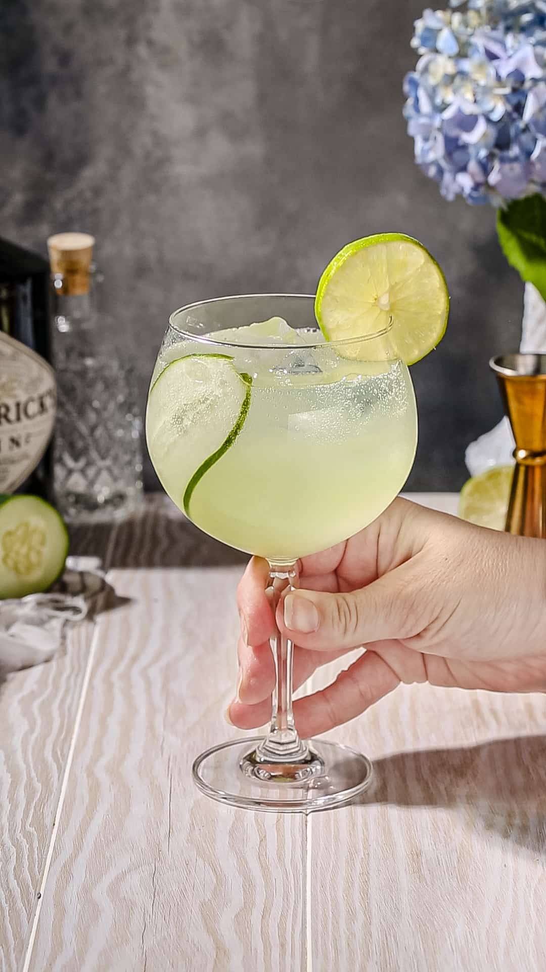 Hand holding the stem of a tall gin and tonic glass filled with a cucumber slice and a light green liquid, with a lime wheel on the rim.