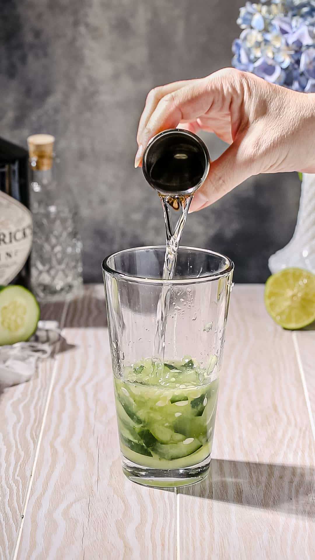 Hand pouring gin from a jigger into a cocktail shaker filled with cucumbers and green liquid.