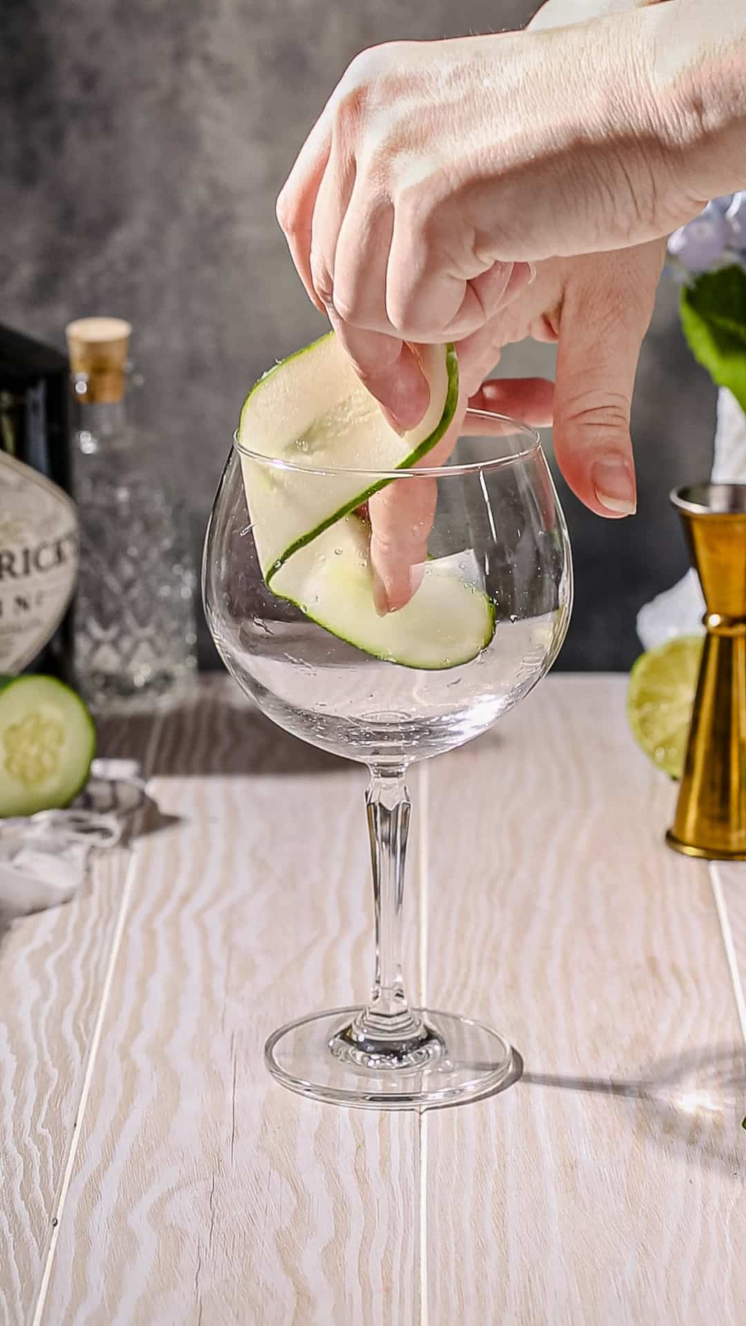 Hands adding a long and flat slice of cucumber to the inside of a tall stemmed cocktail glass.