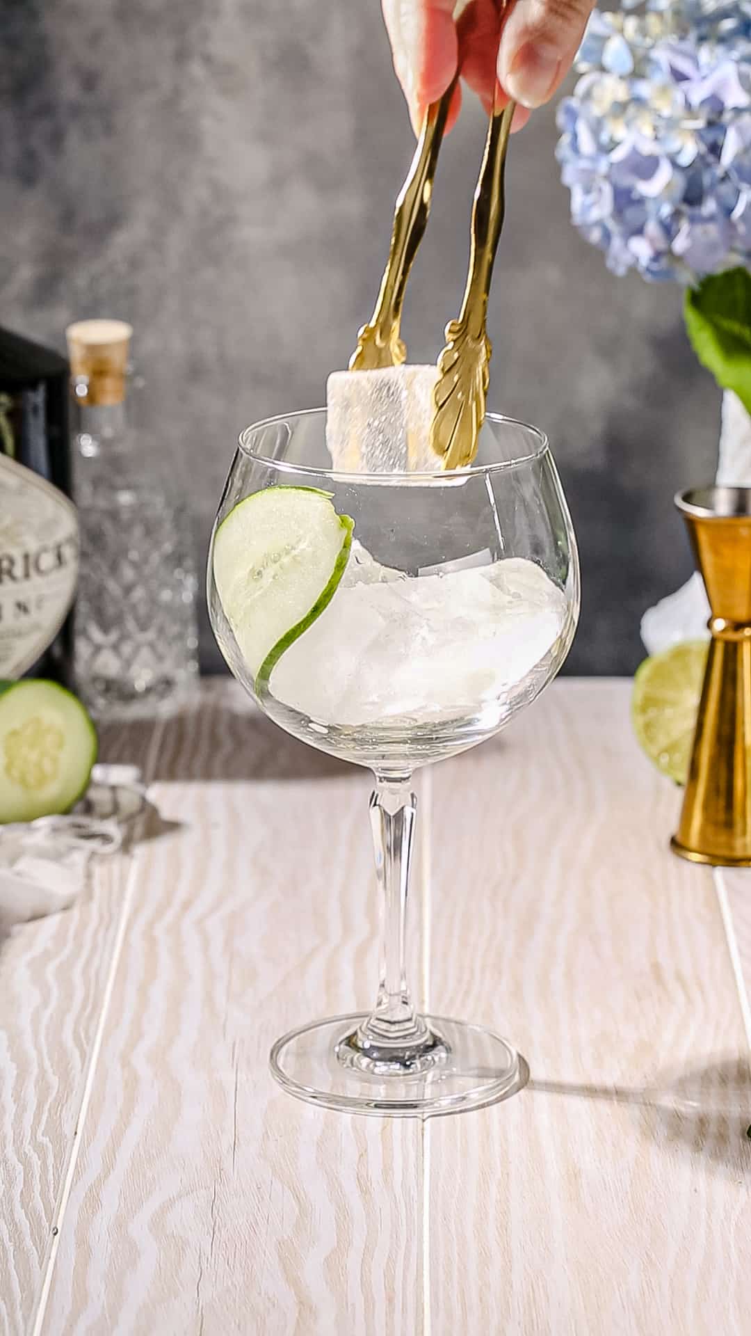 Hands using gold tongs to add a piece of ice to a cocktail glass that has a long thin slice of cucumber inside.