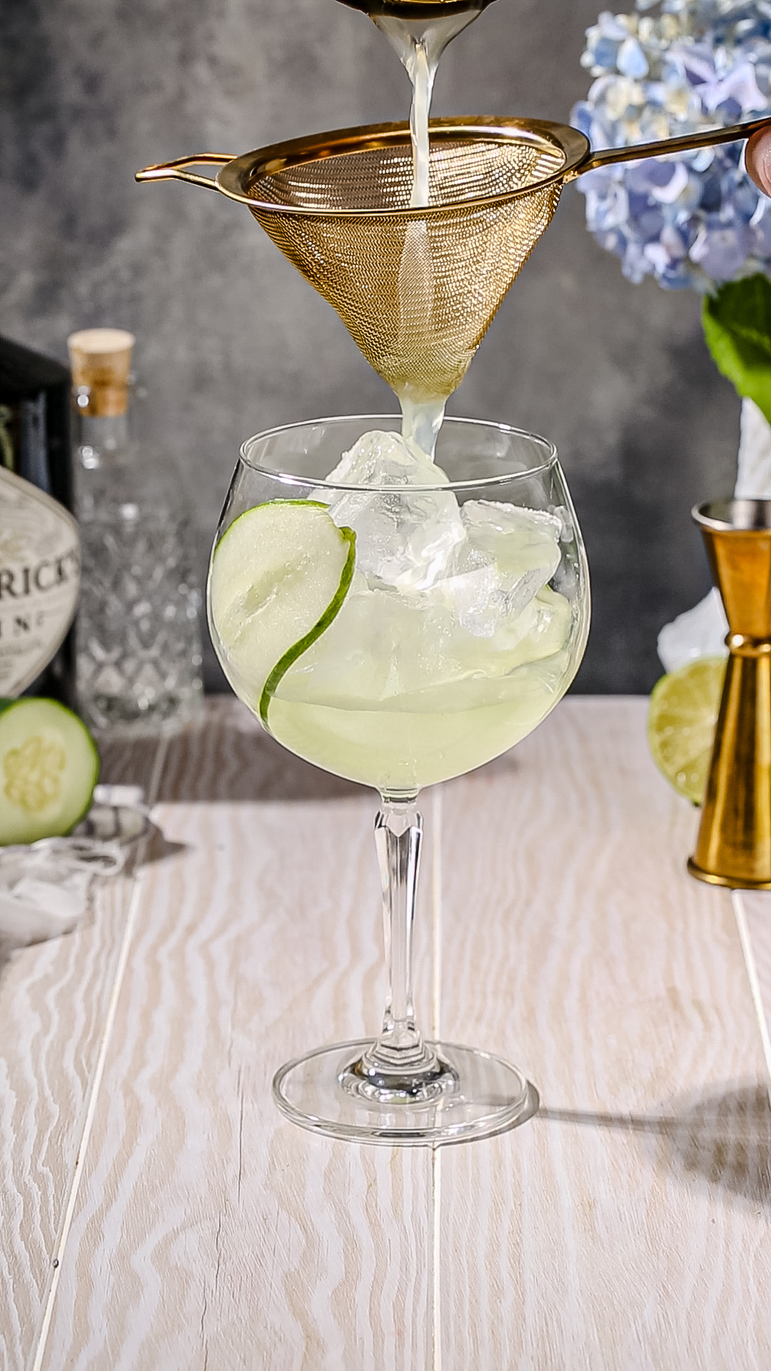 A light green liquid being poured through a gold colored fine mesh strainer into an ice filled glass that also has a cucumber slice in it.