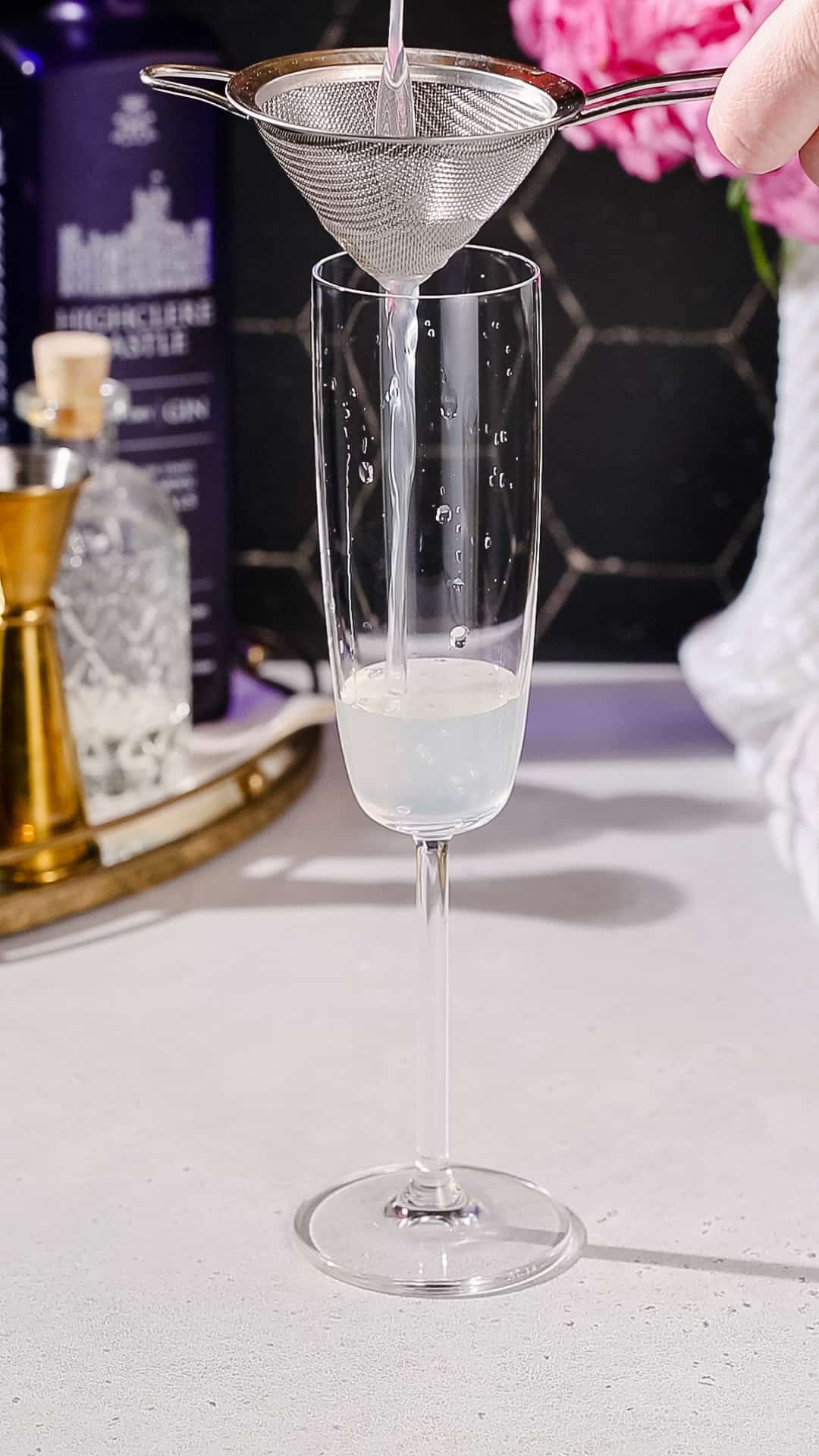 Hand pouring a cocktail into a champagne flute using a fine strainer.