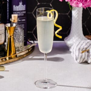 Side view of French 75 cocktail with a long curly lemon peel garnish, and flowers and cocktail ingredients in the background.